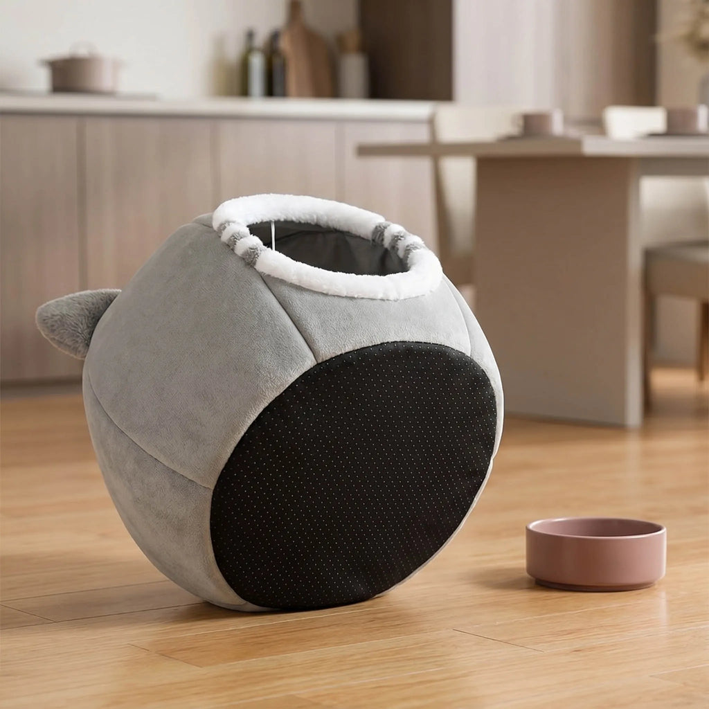 A round plush pet bed with gray fabric, a black textured panel, and a white trim around the opening, shown on a wooden floor in a softly blurred home interior. A small pink bowl is placed beside it.
