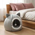 A gray cat-shaped pet bed with ear-like protrusions and a circular opening, placed on a wooden floor beside a bed with pink pillows and white bedding.