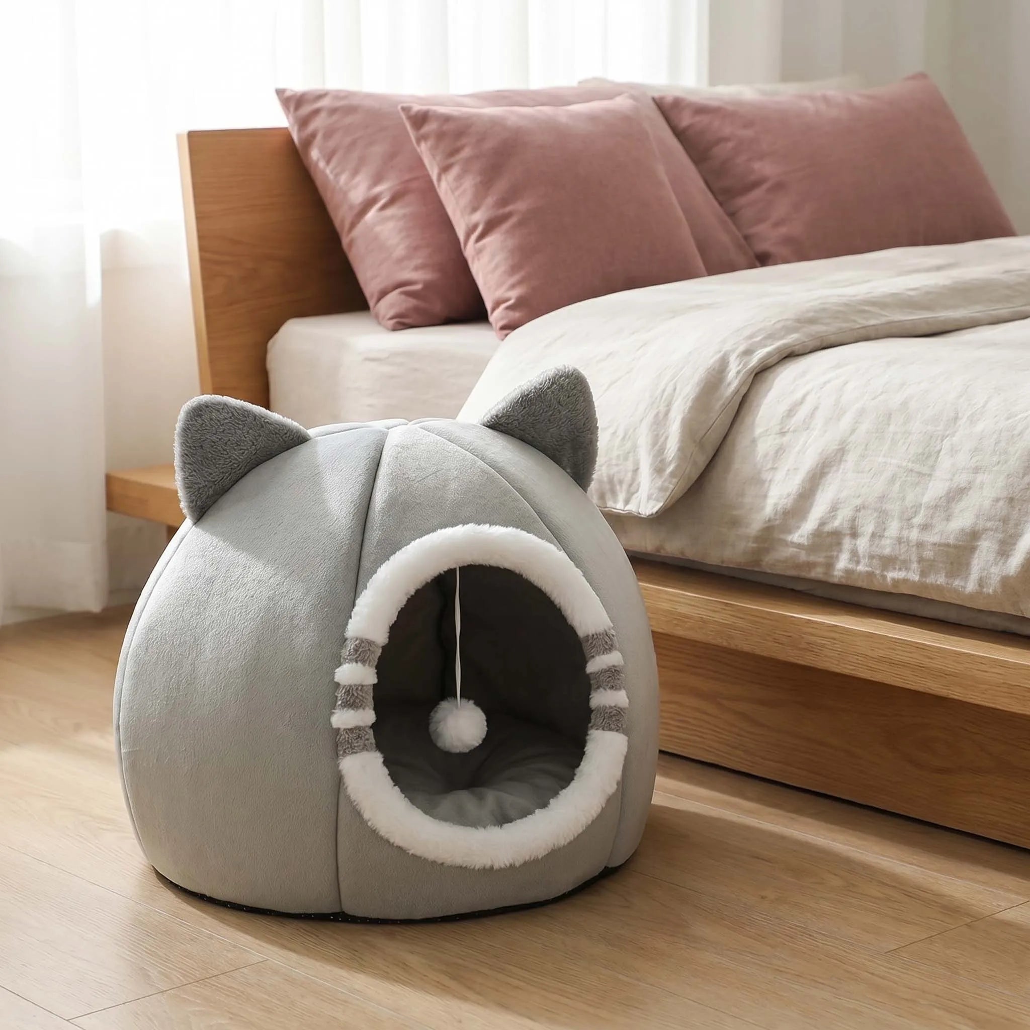A gray cat-shaped pet bed with ear-like protrusions and a circular opening, placed on a wooden floor beside a bed with pink pillows and white bedding.