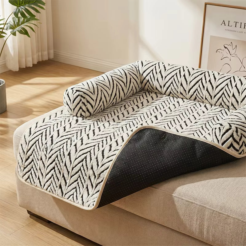 A pet sofa pad with a black and white herringbone pattern and a black underside, shown draped over a beige sofa in a bright, modern living room.