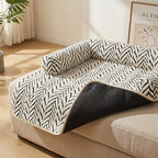 A pet sofa pad with a black and white herringbone pattern and a black underside, shown draped over a beige sofa in a bright, modern living room.