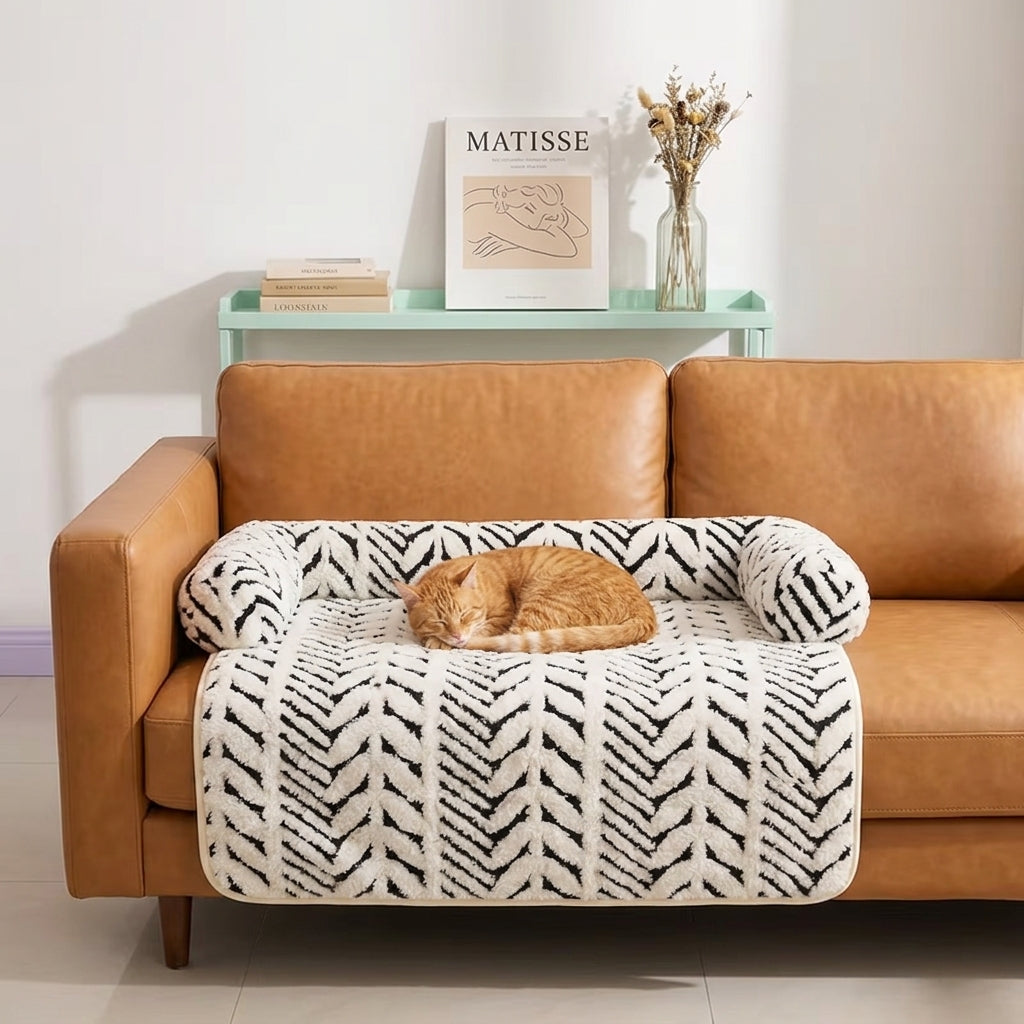 A brown leather sofa in a living room setting with a ginger cat sleeping on a white and black patterned cat bed mat over the seat. A mint green shelf in the background holds a book, a framed print, and a glass vase with dried flowers.