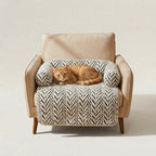 A beige upholstered armchair with wooden legs, styled with a cream and black herringbone-patterned pet bed and a sleeping orange tabby cat resting on top, set against a plain light beige studio background.