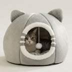 A gray plush cat house with ear-like protrusions, a circular opening lined with white fleece, and a hanging white pom-pom toy. A gray tabby cat is sitting inside the house against a clean white background.