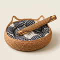 A round woven straw pet bed with a navy and white wave-patterned cushion, featuring a rolled-up mat.
