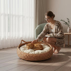 A woman crouches beside a woven round pet bed on a wood floor, gently petting a sleeping orange cat resting on a cream cushion inside the bed. The room has sheer white curtains, a light green chair, and a small side table in the background.