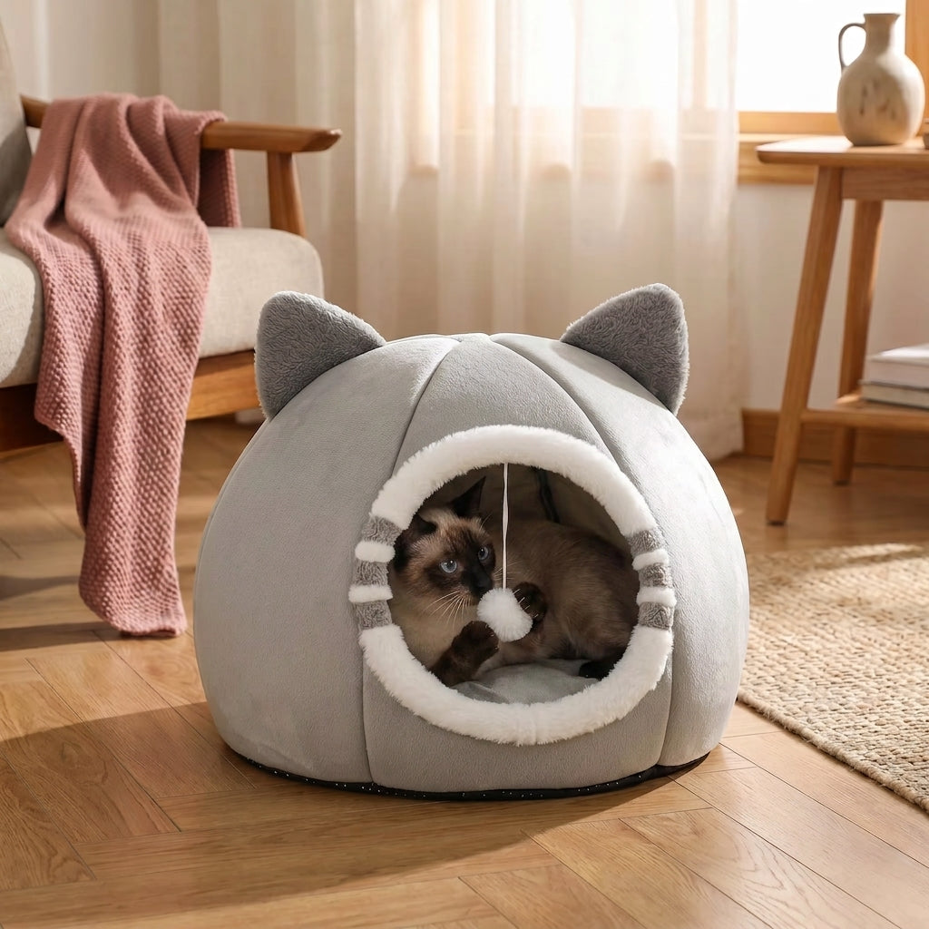 A gray cat bed shaped like a cat head with ears, shown on a wooden floor in a living room. A cat is lying inside the opening, playing with a hanging toy.