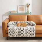 An orange upholstered sofa with wooden legs, styled with a white and black herringbone-patterned pet bed and a sleeping orange tabby cat on top, set against a white wall and a pastel-colored table. The table has some books and a glass flower vase on top of it.